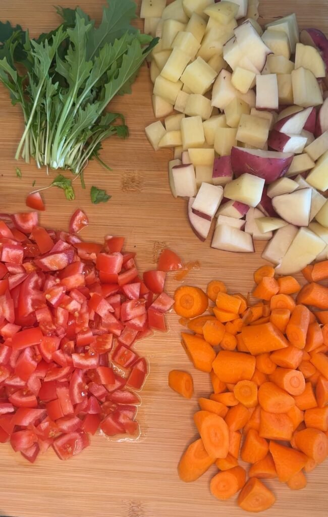 diced potato, tomatoes, carrots, and whole greens on a wood cutting board 